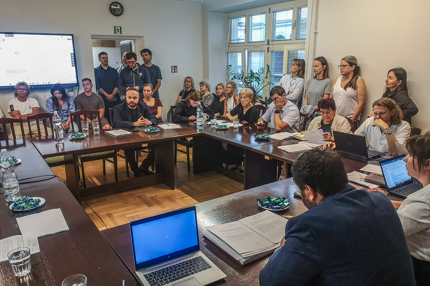 zdjęcie z sesji - radni siedzący przy stołach ustawionych w duży kwadrat w tle liczni mieszkańcy