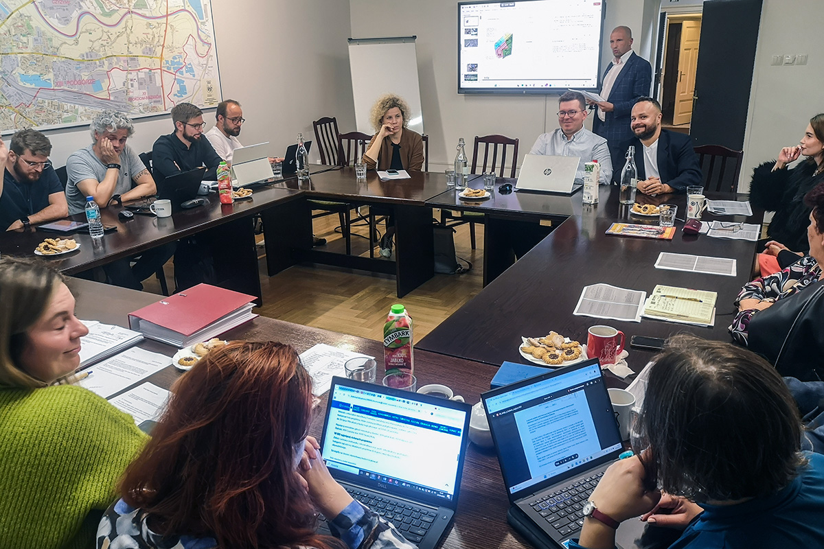 zdjęcie z sesji - radni siedzący przy stołach ustawionych w duży kwadrat w tle liczni mieszkańcy