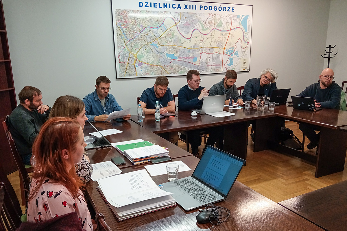 zdjęcie z sesji - radni siedzący przy stołach ustawionych w duży kwadrat w tle liczni mieszkańcy